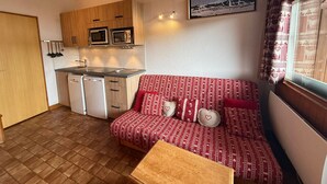 A compact kitchen with wooden cabinets, a microwave, and a dishwasher. A red patterned sofa with heart-shaped pillows is placed against the wall, and a small wooden table is in front of it. - Les Saisies Apt, 4 pers, south balcony, near slopes (Les Saisies)