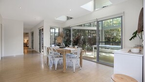 A spacious dining area with a wooden table and white chairs, large sliding glass doors leading to a garden, and a high ceiling with skylights. - The Sanctuary - a tranquil hidden oasis (Hawks Nest)