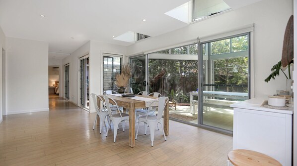 A spacious dining area with a wooden table and white chairs, large sliding glass doors leading to a garden, and a high ceiling with skylights. - The Sanctuary - a tranquil hidden oasis (Hawks Nest)
