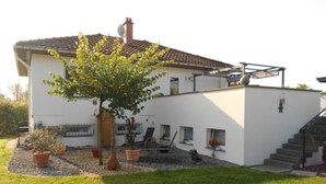 Une maison à deux étages, avec une façade blanche, un toit en tuiles et un petit jardin agrémenté de plantes en pot et d’un arbre.