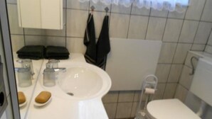 Une petite salle de bain avec un lavabo blanc, des toilettes et un espace douche. Deux serviettes noires sont accrochées à des crochets au-dessus du lavabo.