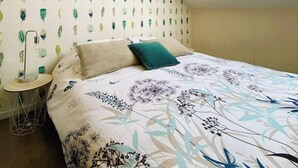 Une chambre avec un plafond en pente, un puits de lumière, un lit avec une literie à motifs floraux, une lampe de chevet et un mur orné de motifs de feuilles.