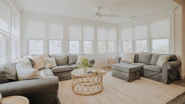 Un salon spacieux doté d’une grande fenêtre, d’un ventilateur de plafond et d’un canapé d’angle gris.