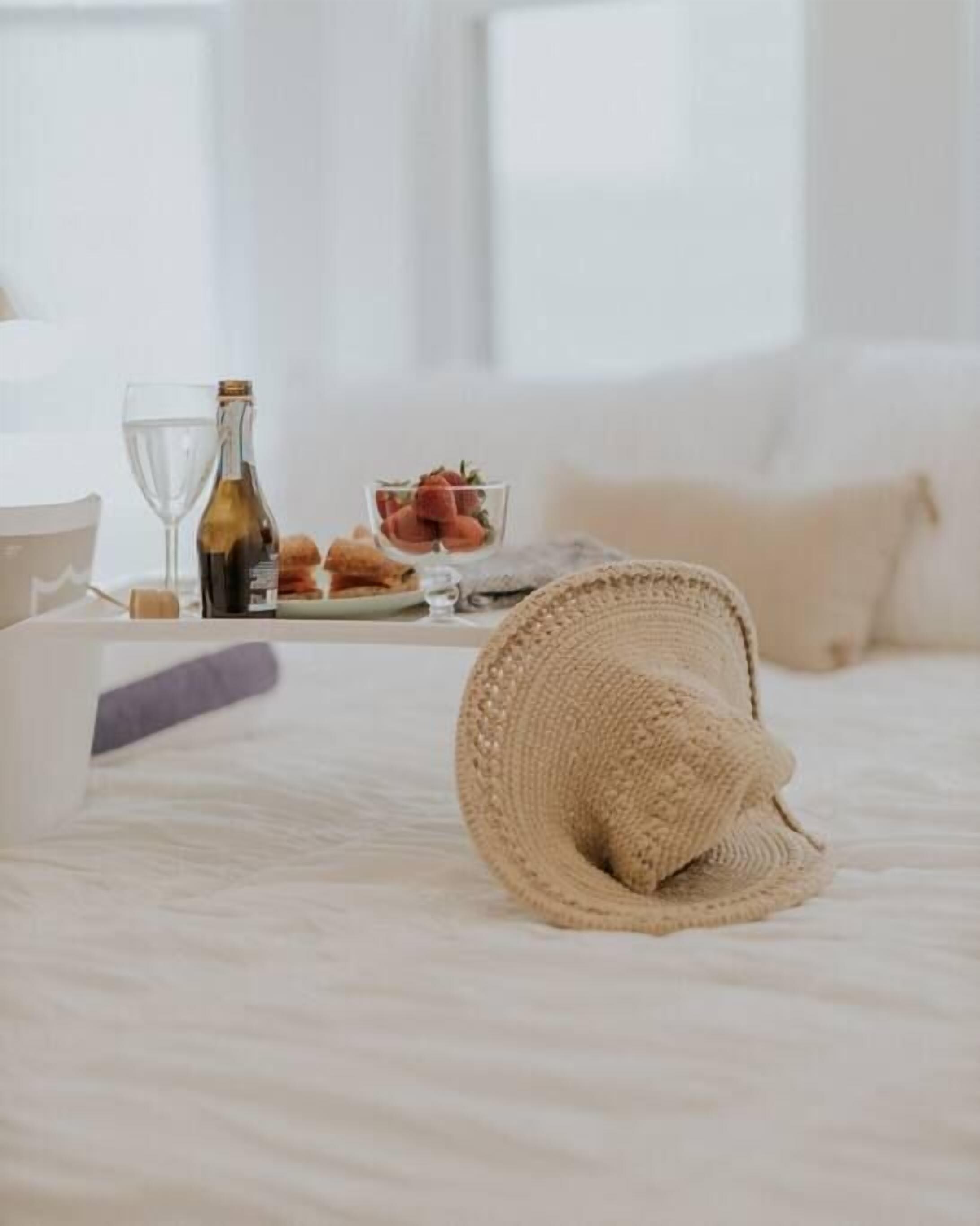 A neatly made bed with a rolled-up towel, a bottle of wine, and a bowl of strawberries on a tray.