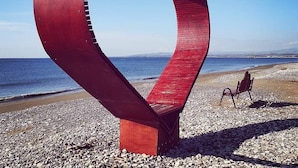 Une sculpture métallique en forme de cœur, placée sur une plage de galets, avec une chaise et un banc à proximité.