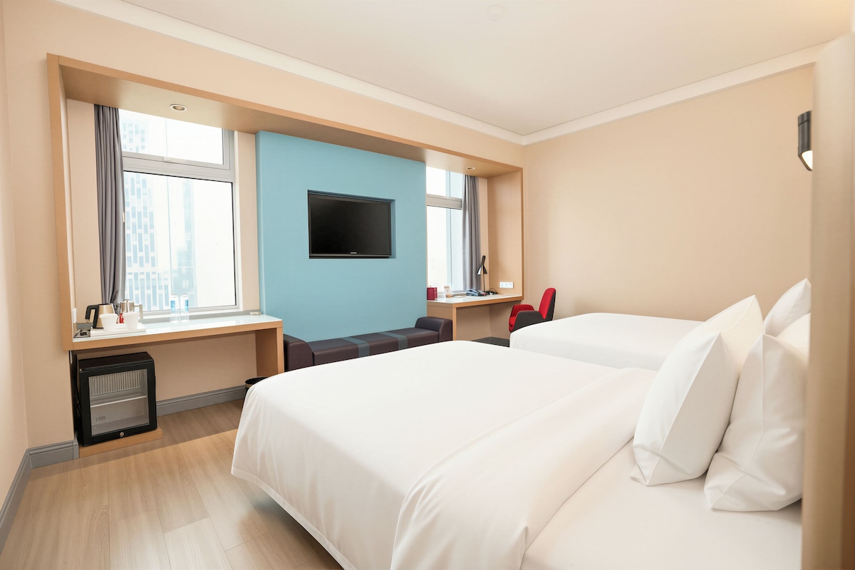 A hotel room with two beds, a desk, a TV, and a window with curtains.