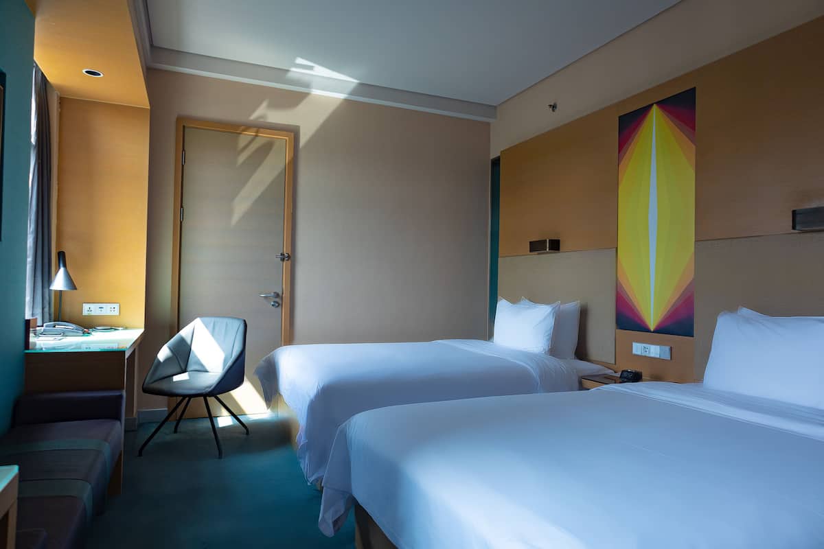 A hotel room with two beds, a desk, a chair, and a colorful wall art piece.