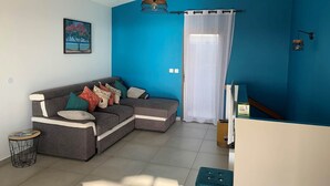 A living room with a grey sectional sofa, a small round side table, a ceiling fan, a wall-mounted light fixture, and a window with a sheer curtain. - Belle Villa Avec Piscine à 200m de la Plage ! (Saint-Leu)