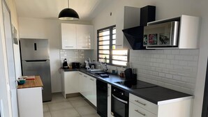 A modern kitchen with white cabinets, a stainless steel refrigerator, a black countertop, and a window providing natural light. - Belle Villa Avec Piscine à 200m de la Plage ! (Saint-Leu)