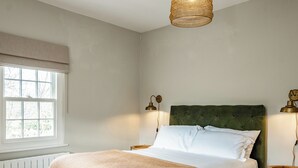 Ein Schlafzimmer mit einem grünen, gepolsterten Kopfteil, einer weißen Bettdecke und einer braunen Tagesdecke. Ein Fenster mit weißen Jalousien, ein Heizkörper darunter und zwei Nachttische mit Holzoberfläche, jeweils mit einer Nachttischlampe.