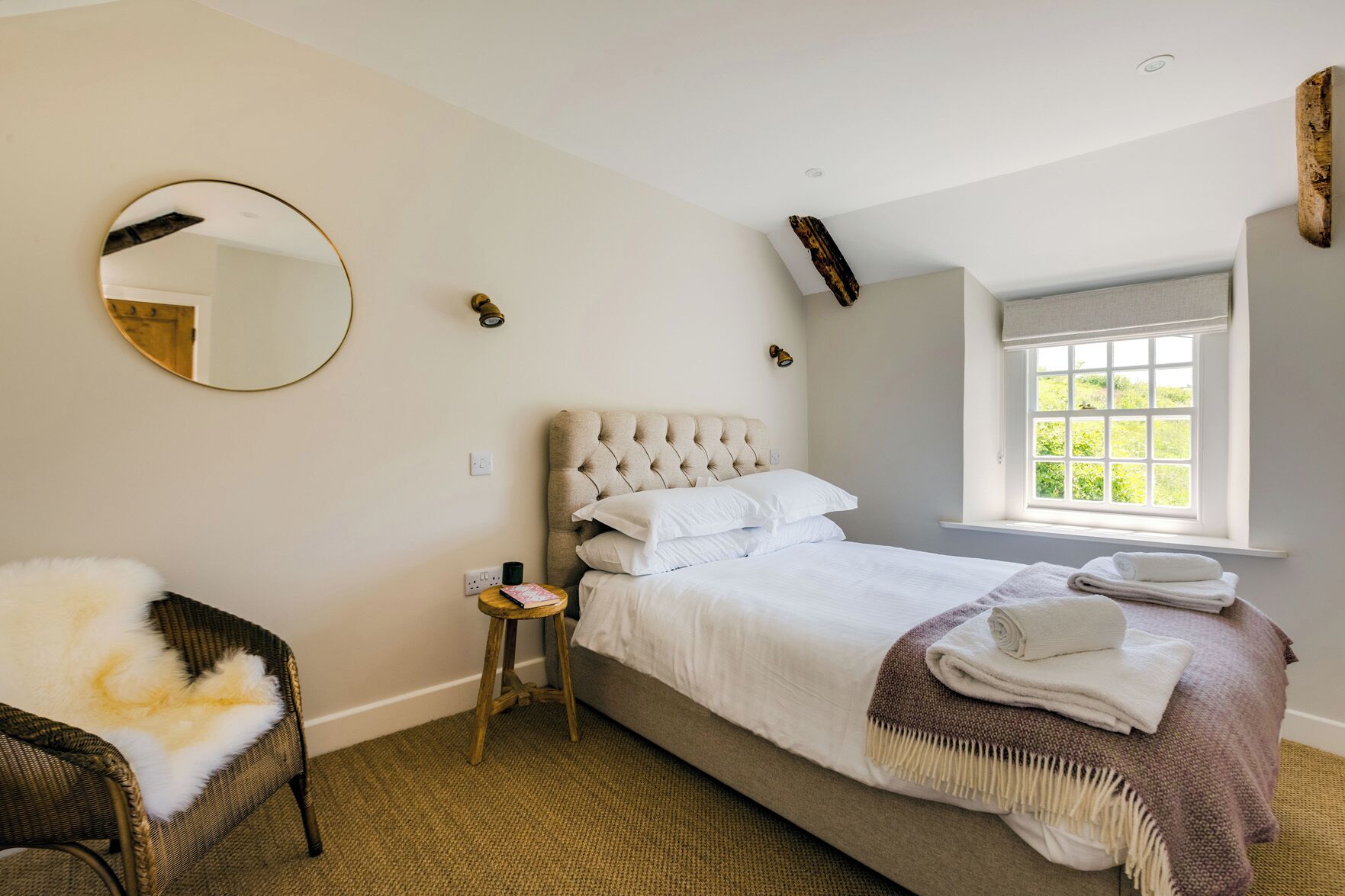 A bedroom with a large bed, a round mirror, a small stool, and a window with a view of greenery.