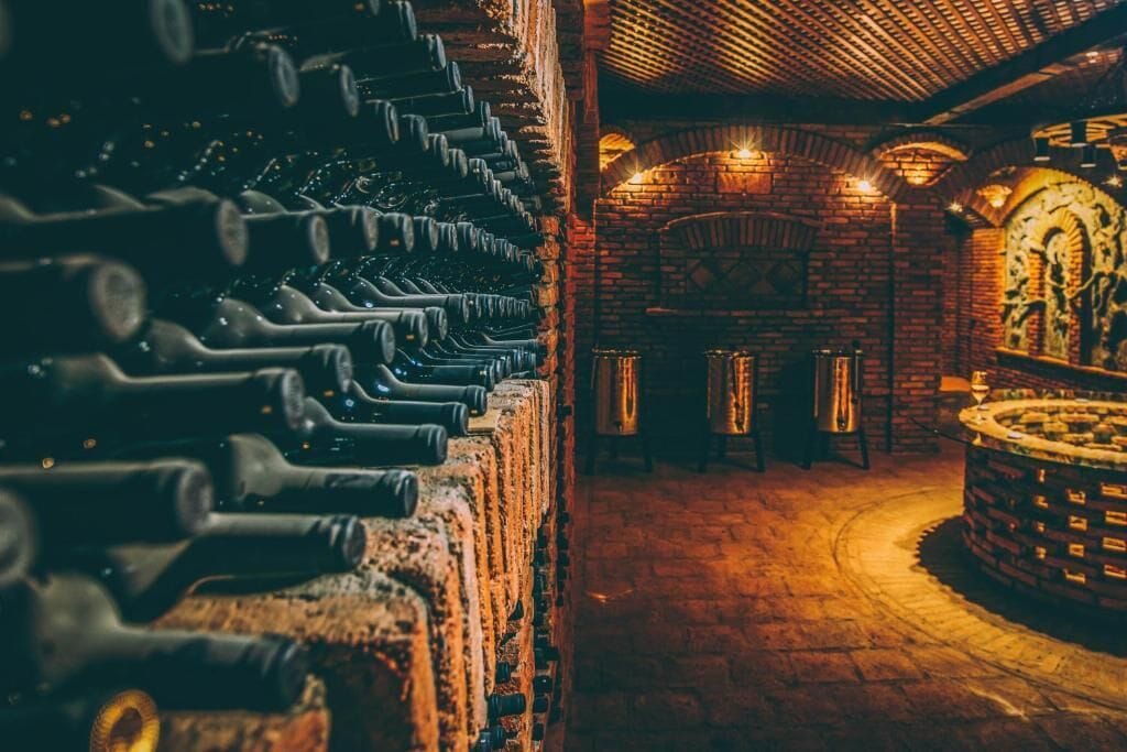 Una cava con filas de botellas y barriles de vino, paredes de ladrillo y una estructura circular de piedra.