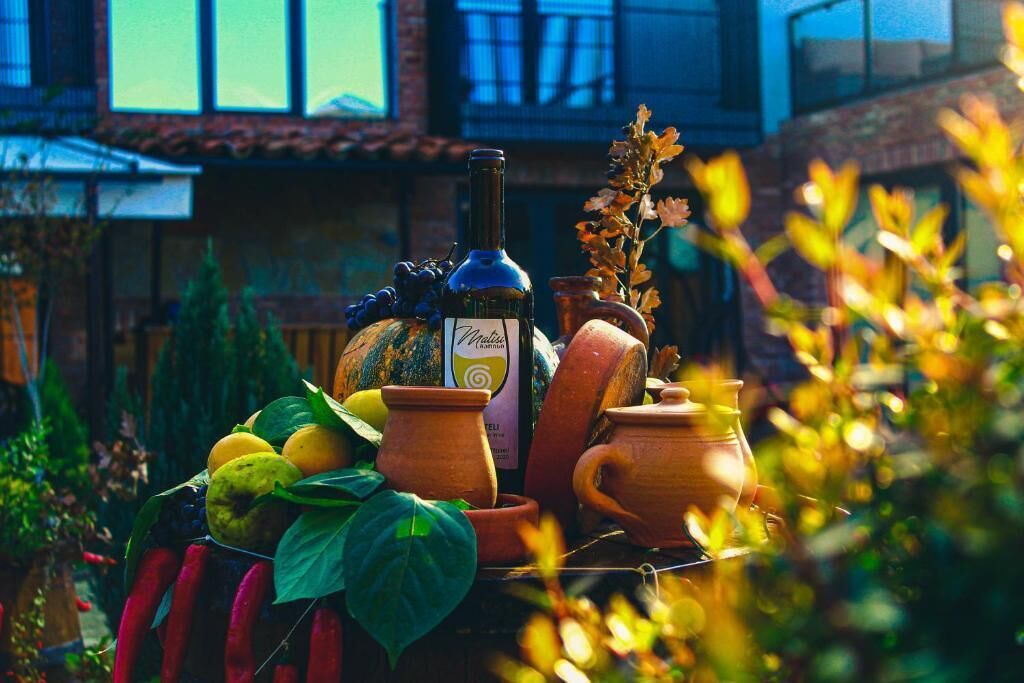 Ein Tisch mit einer Flasche, Früchten und Keramik im Freien, mit einem Gebäude im Hintergrund.