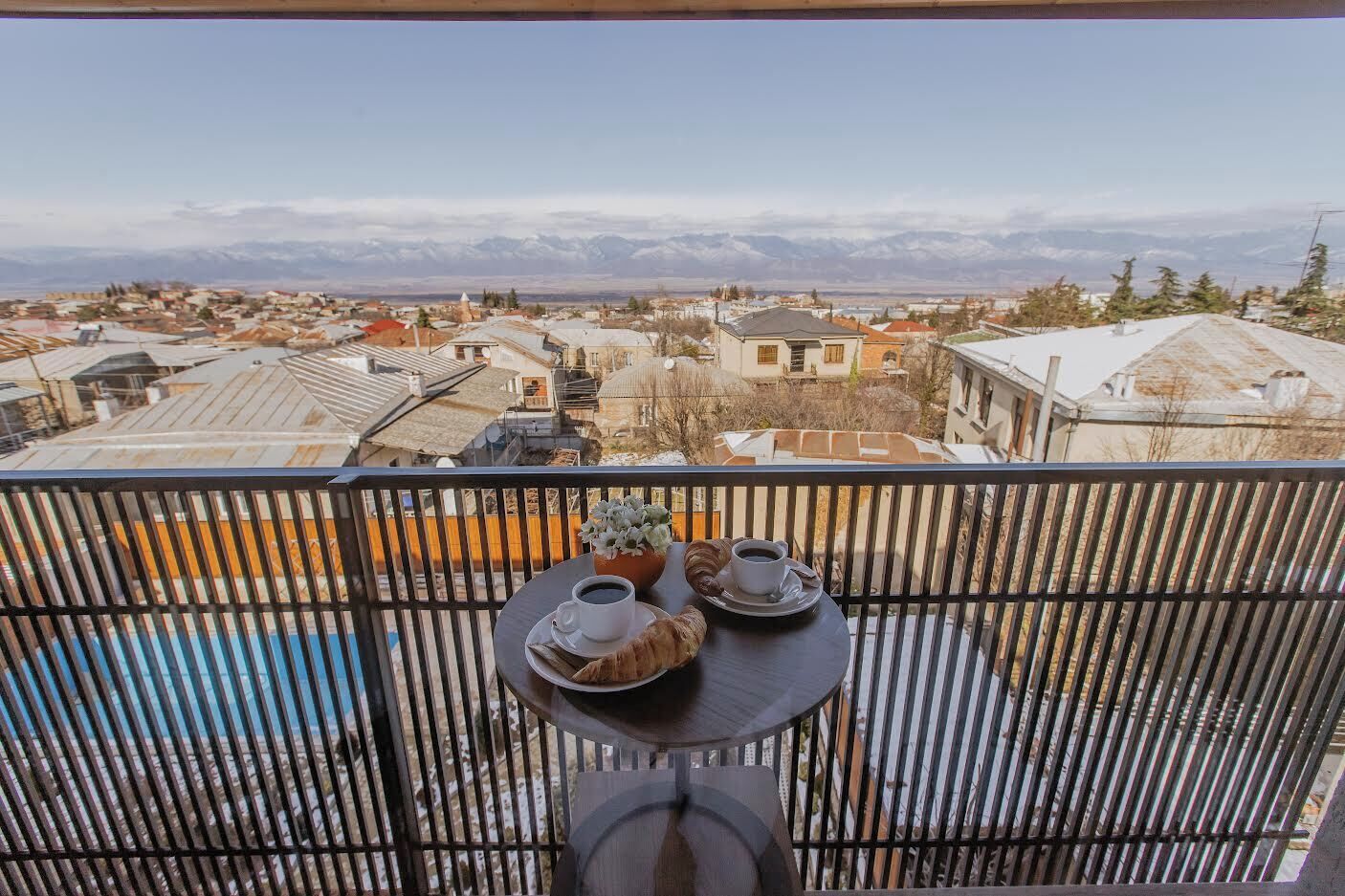 A balcony with a table set for two, overlooking a residential area with snow-covered roofs and distant mountains.