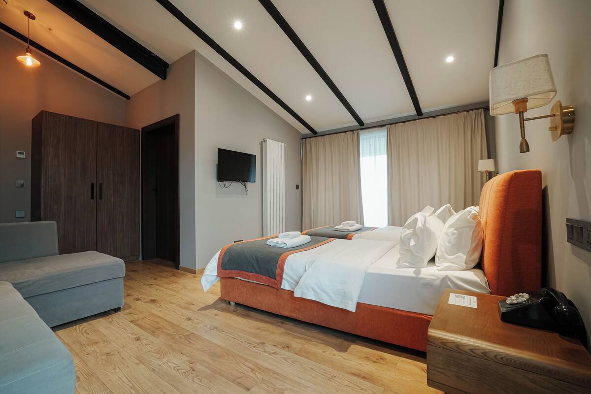 Una habitación de hotel moderna con una cama grande, un sofá, piso de madera, un televisor y una mesita de noche con lámpara.