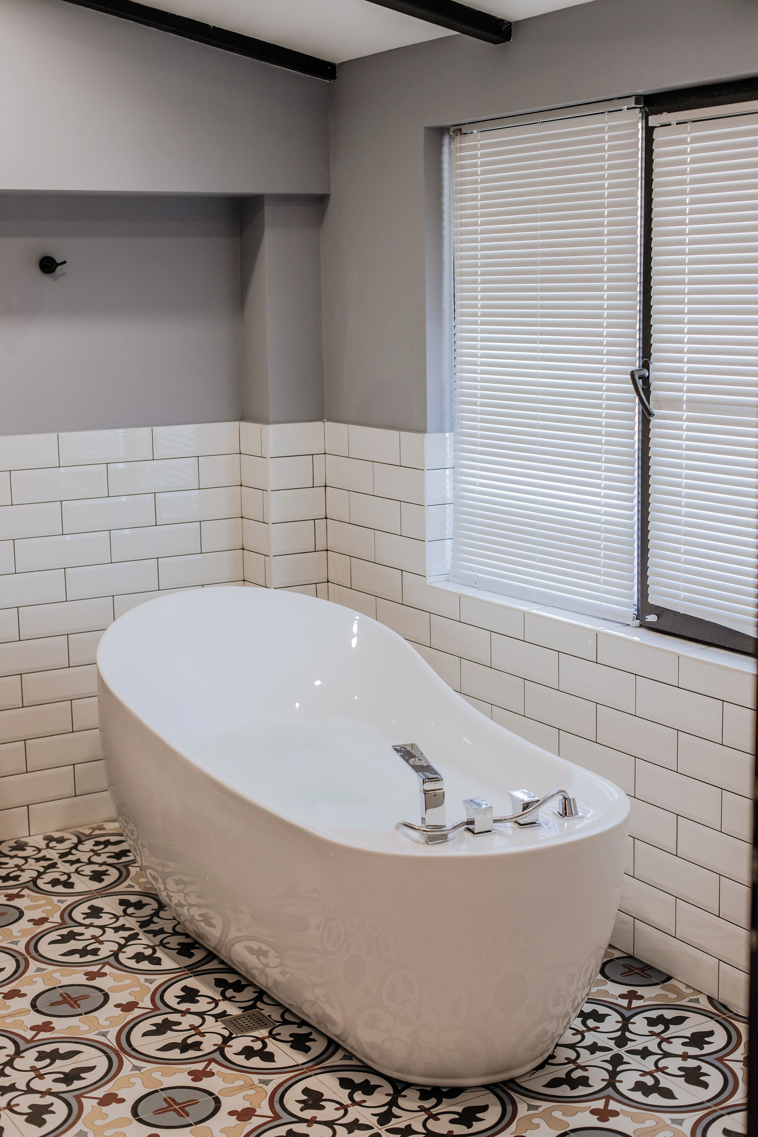 Un baño moderno con una bañera independiente, paredes de azulejos blancos y una ventana con persianas.