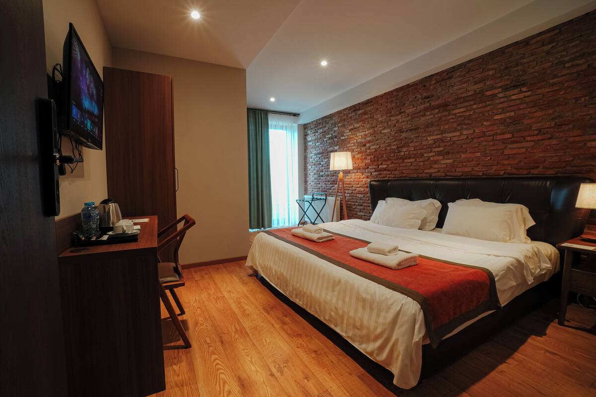 Habitación de hotel con pared de ladrillo, cama con ropa blanca, escritorio con hervidor y televisor montado en la pared.