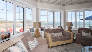 Een ruime woonkamer met grote ramen met uitzicht op een strand, ingericht met banken en een salontafel.
