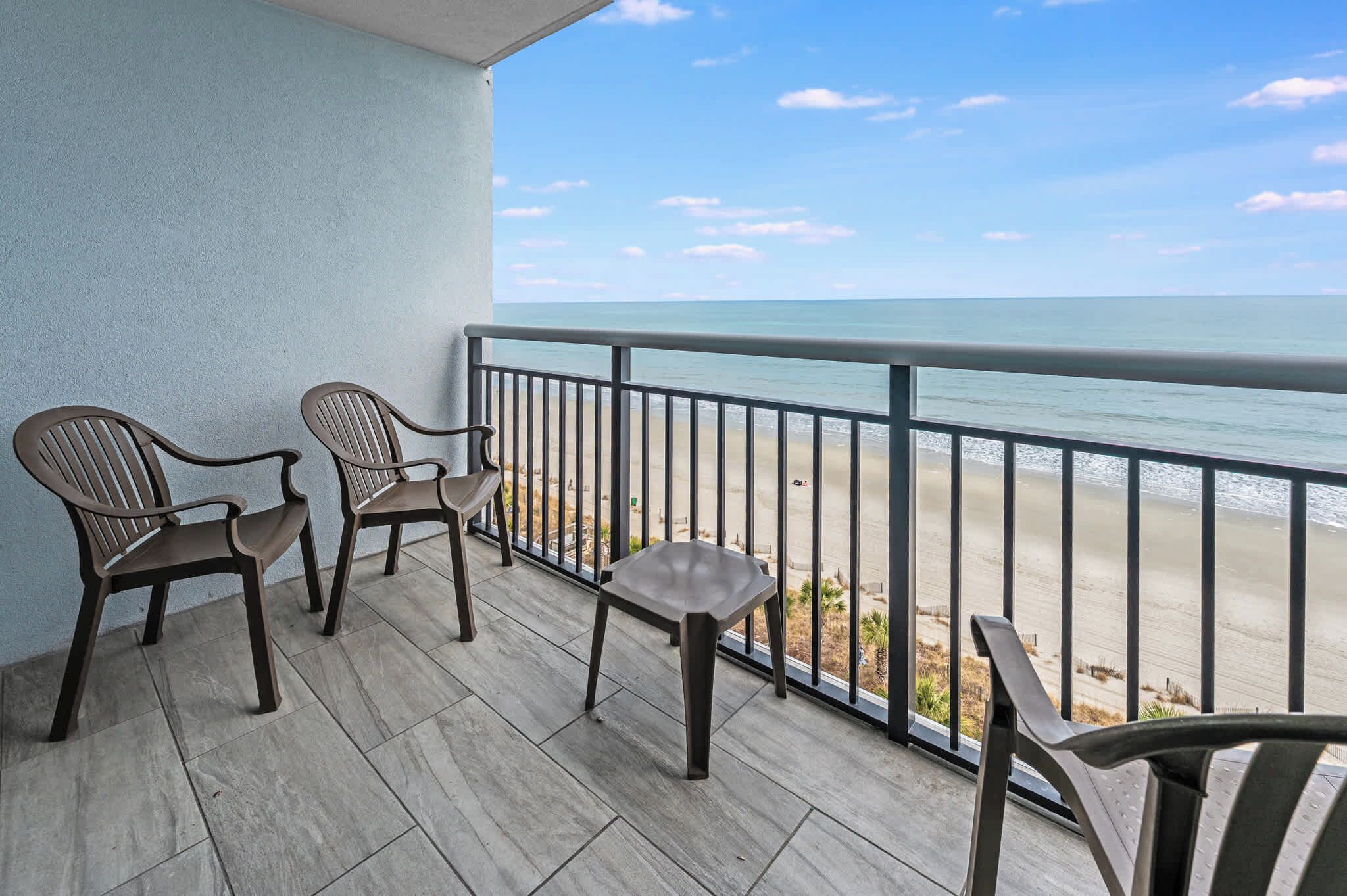 Un balcon avec deux fauteuils en osier et une petite table, donnant sur une plage sous un ciel dégagé.
