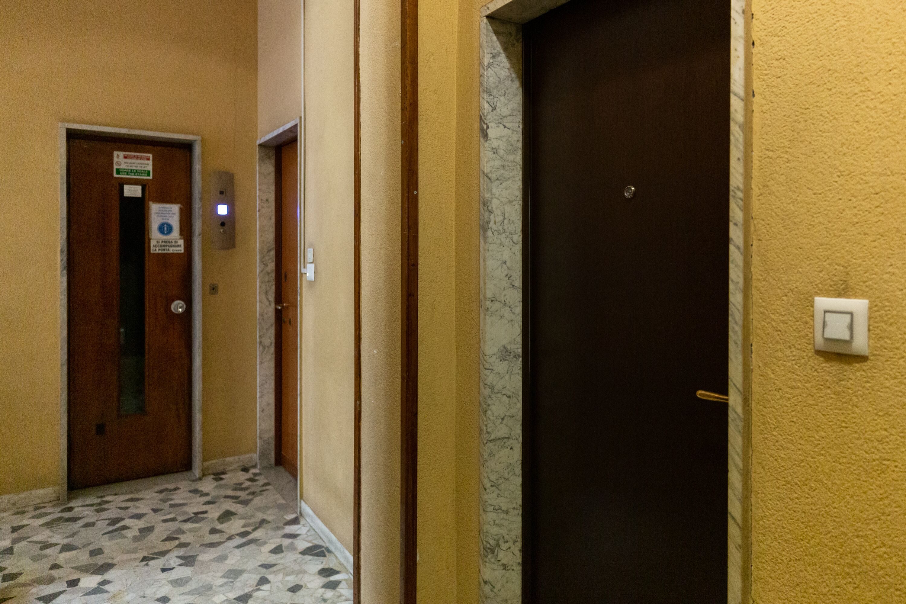 A hallway with a marble floor, a wooden door, and a wall-mounted light switch.
