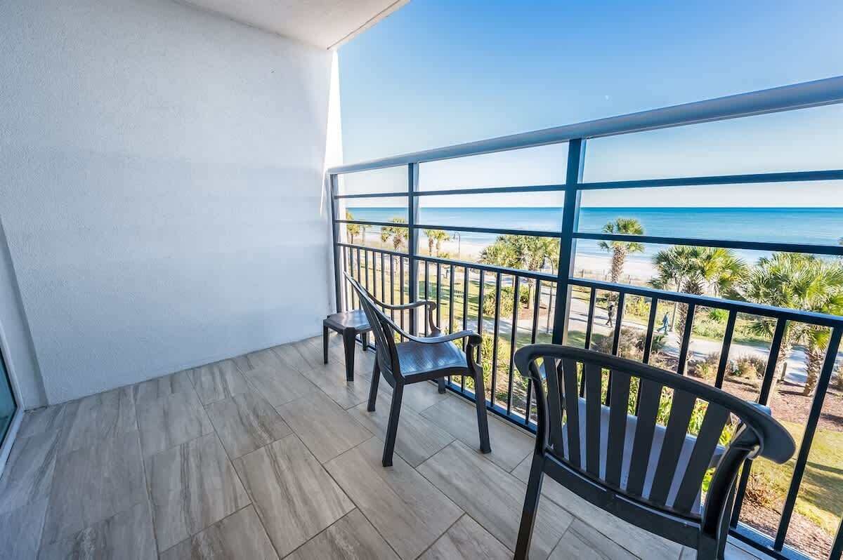 Een balkon met uitzicht op het strand, met twee zwarte stoelen en een tafel.