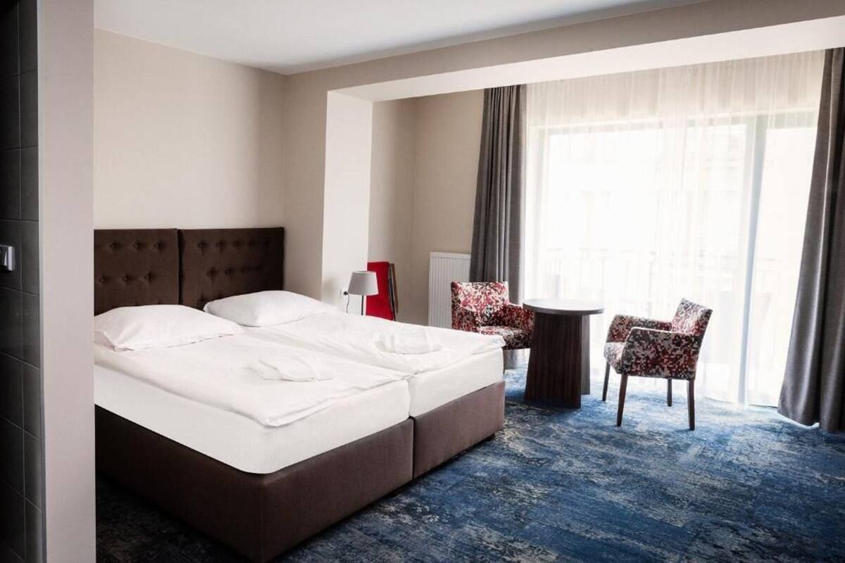 A hotel room with a large bed, two chairs, a small table, and a window with curtains.