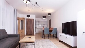Uma sala de estar moderna com sofá, mesa de centro e rack para TV. Há uma área de jantar com cadeiras e uma mesa. A cozinha fica ao fundo.