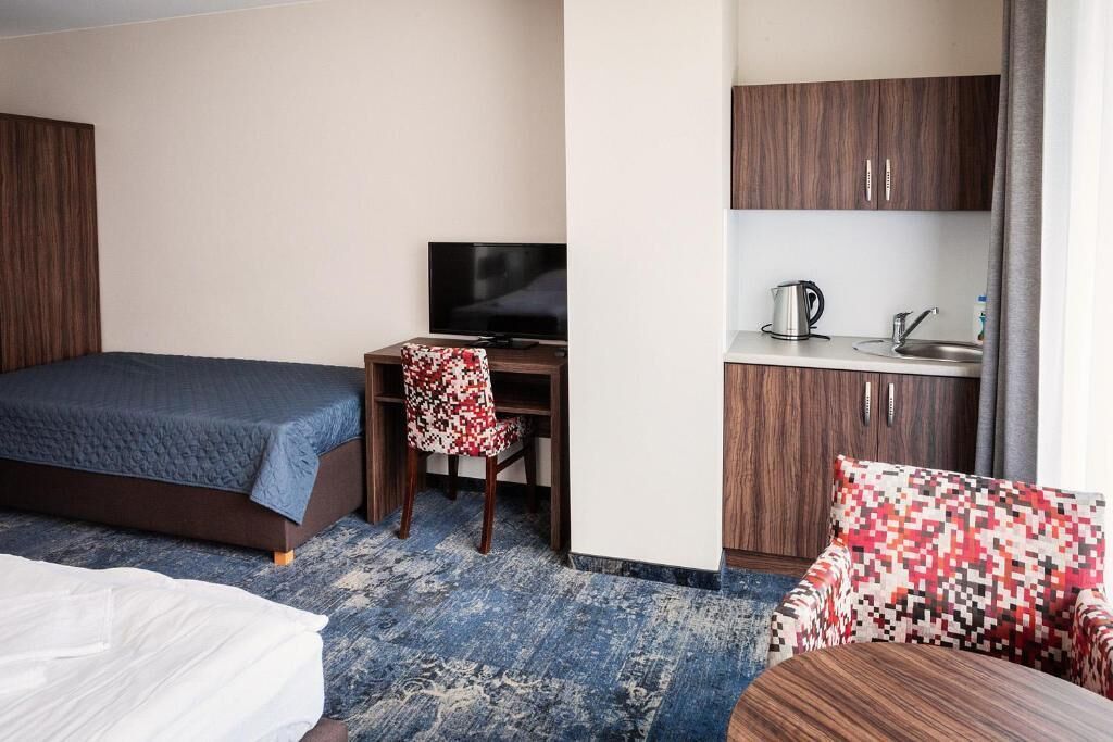 Une chambre d’hôtel compacte comprenant un lit, un bureau avec une télévision, une petite kitchenette et un fauteuil à motifs.