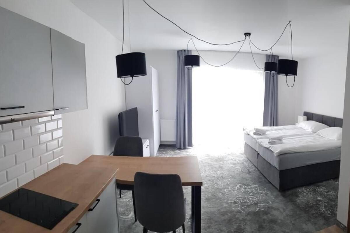 A modern hotel room with a bed, a desk with a chair, a television, and a kitchen area with a fridge and cabinets.