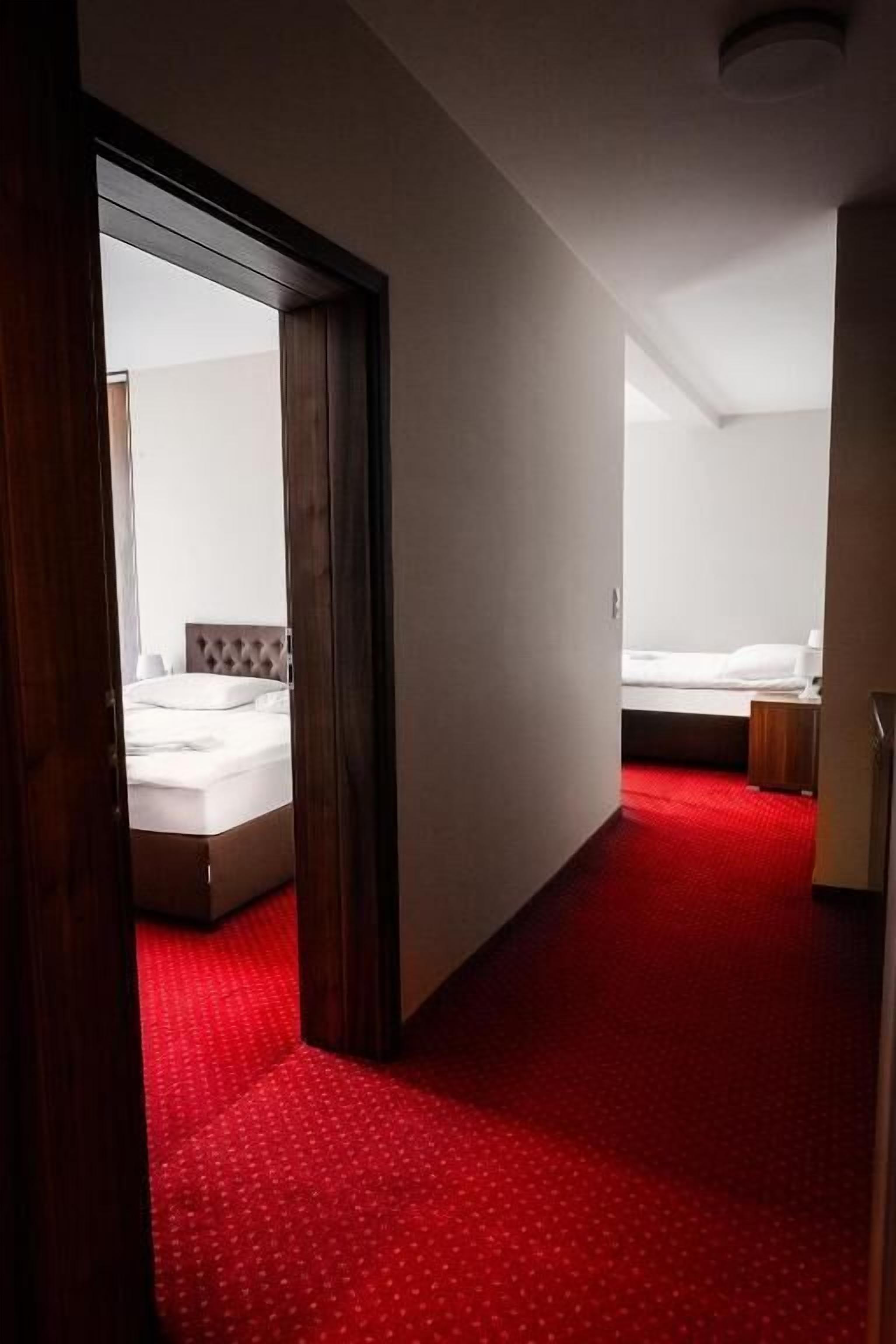 A hotel corridor with a red carpet leading to a room with a bed and a nightstand.