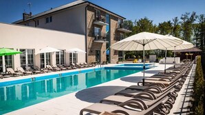 Uma área de piscina com espreguiçadeiras e guarda-sóis, um edifício com varandas, e um céu claro.