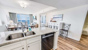 A modern kitchen with a central island, stainless steel sink, and white cabinetry. - King Suite 3BR,Direct Oceanfront Views, Remodeled! (Myrtle Beach)