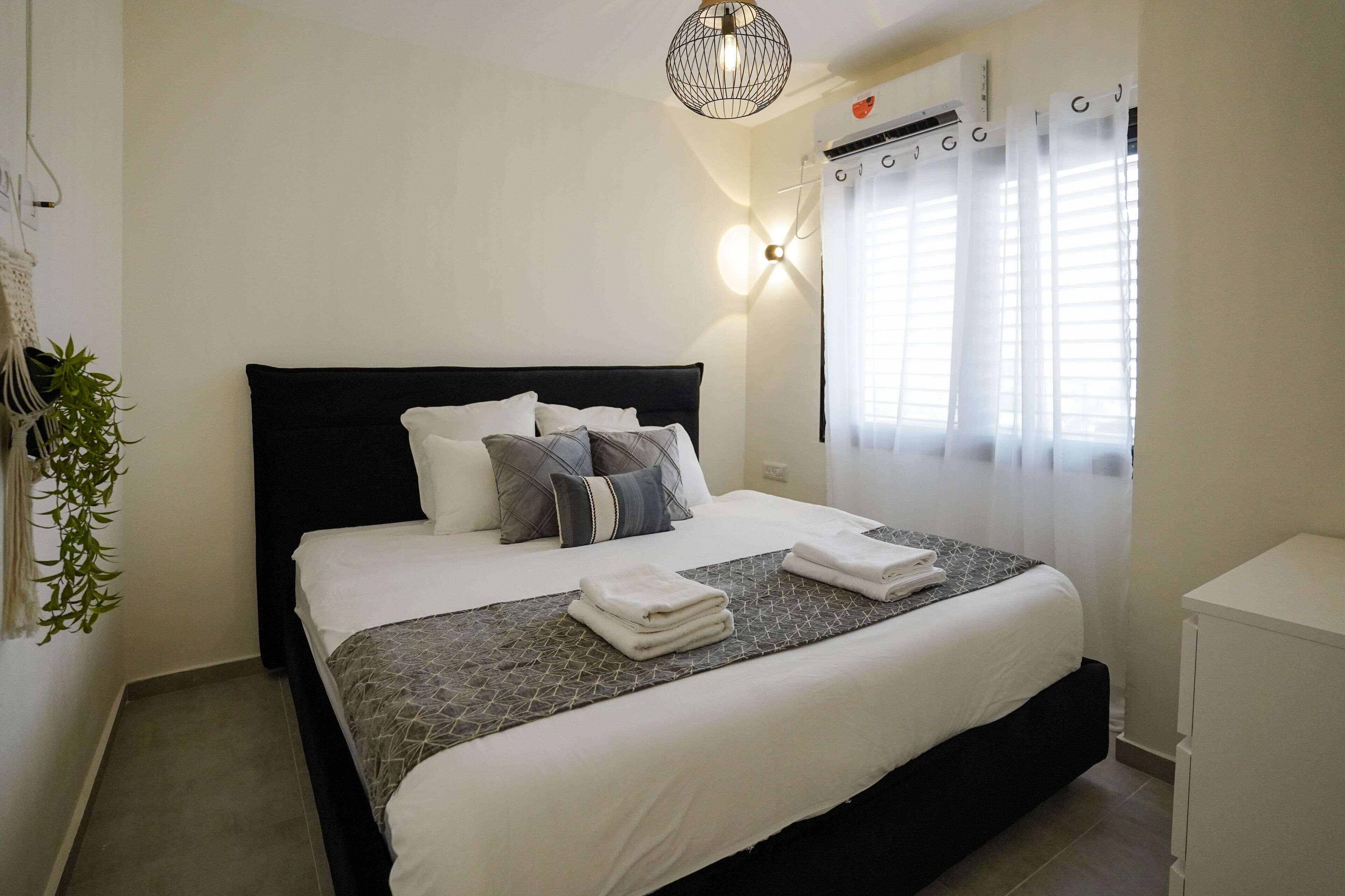 Una cama bien hecha con cabecera negra, ropa de cama blanca y una manta gris estampada. Se ve una unidad de aire acondicionado de pared y una planta colgante a la izquierda.