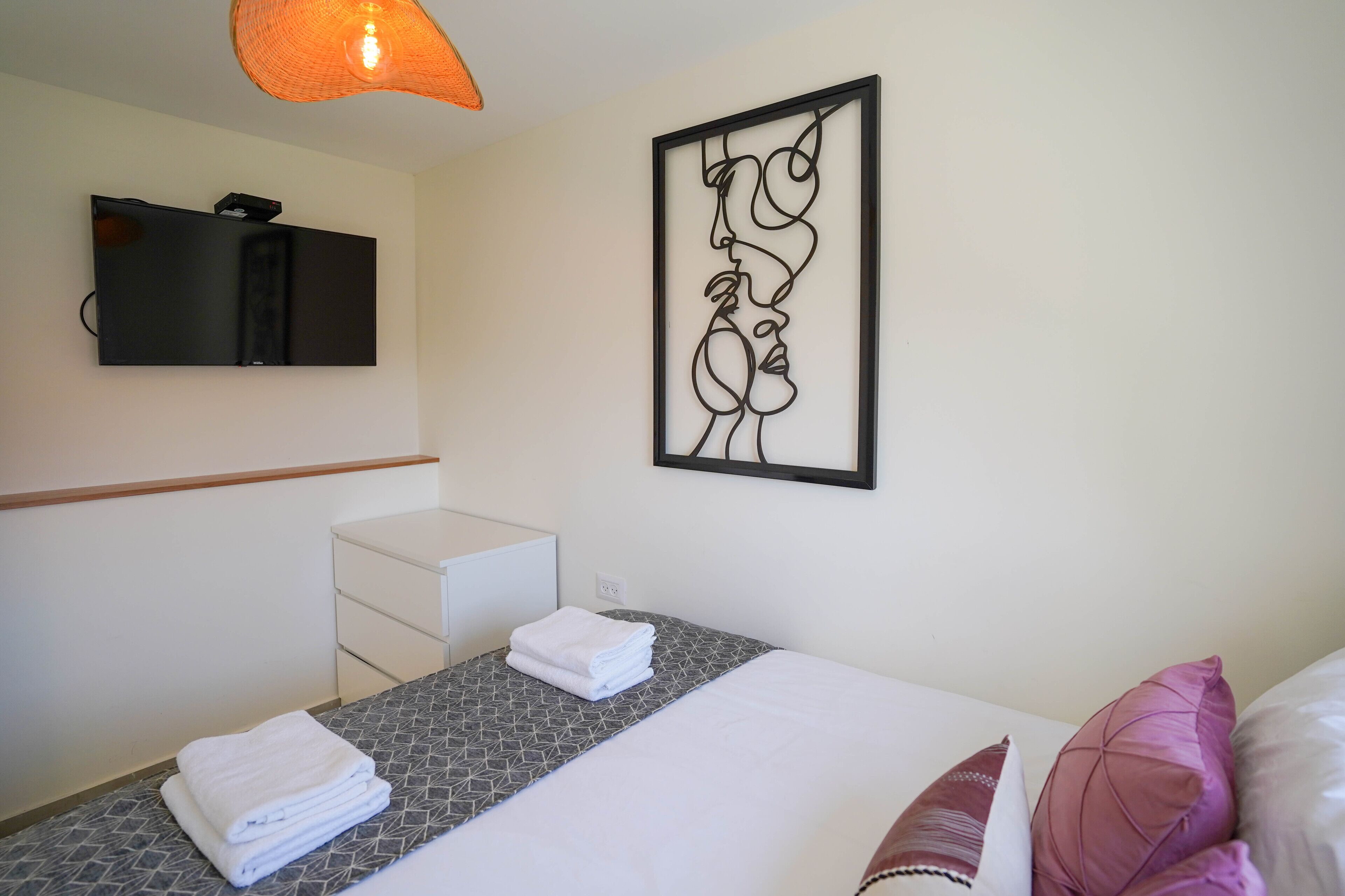 Quarto de hotel com cama, TV fixada na parede, uma obra de arte na parede e mesa de cabeceira com toalha dobrada.