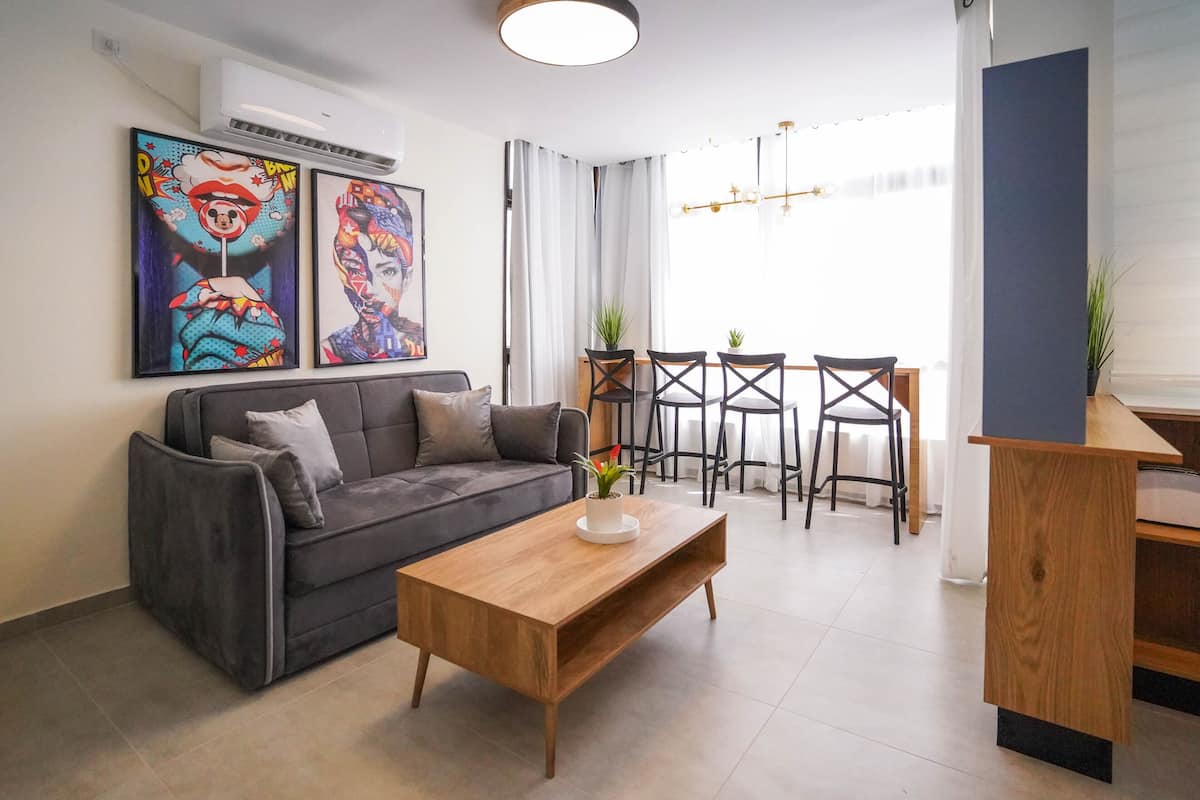 A modern living room with a grey sofa, wooden coffee table, and a dining area with a table and chairs. There are two paintings on the wall and a potted plant on the coffee table.