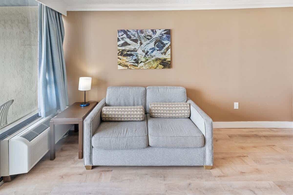 Uma sala de estar com um sofá cinzento, uma mesinha de apoio, um abajur e um quadro na parede.