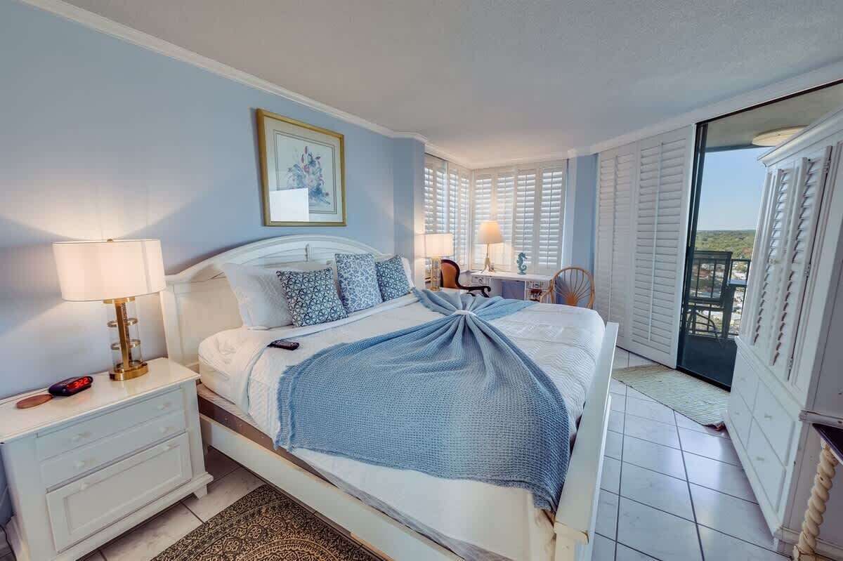 A bedroom with a bed, bedside table, lamp, and a view of the outdoors through a balcony.