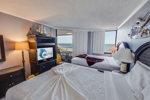 Une chambre d’hôtel avec deux lits, un téléviseur diffusant des publicités pour « BIG FISH Rentals », un balcon avec vue sur la mer et un bateau décoratif accroché au mur.