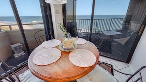 Et rundt træbord med fire servietringe, en dekorativ tepotte og en vase med blomster på en balkon med udsigt over havet.