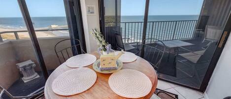 A round wooden table with four placemats, a decorative teapot, and a vase with flowers on a balcony overlooking the ocean.