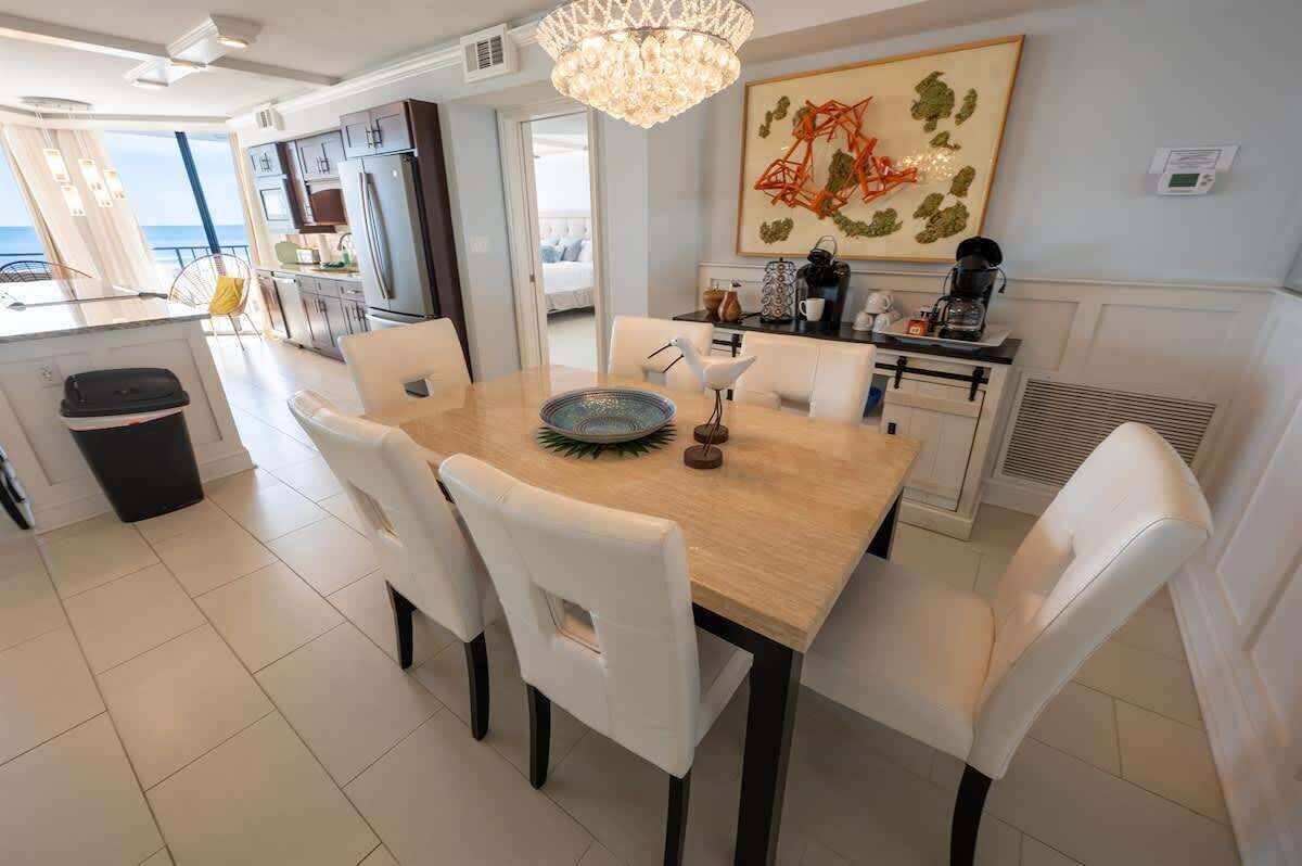 A modern kitchen with white cabinetry, a dining table with white chairs, and a view to the sea.