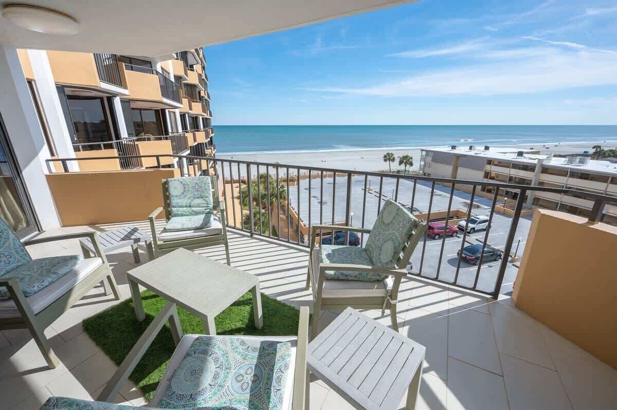 A balcony with a view of the beach, outdoor furniture, and a parking area in the background.