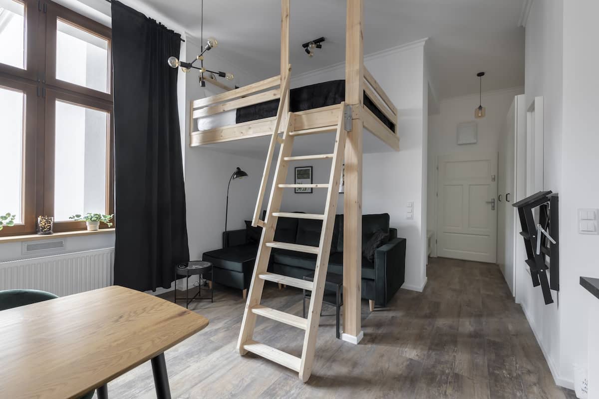 Ein Zimmer mit Etagenbett, Holzleiter, Schreibtisch, Stuhl, Stehlampe und einem Fenster mit Vorhängen.