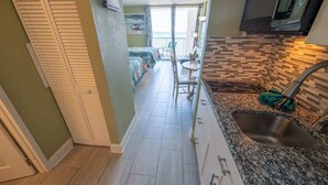 A narrow hallway with a kitchen area featuring a granite countertop and a sink. A bed with a colorful bedspread is visible in the background. - NEW Ocean Annie's Oceanview Studio - Heated Pools! (Myrtle Beach)