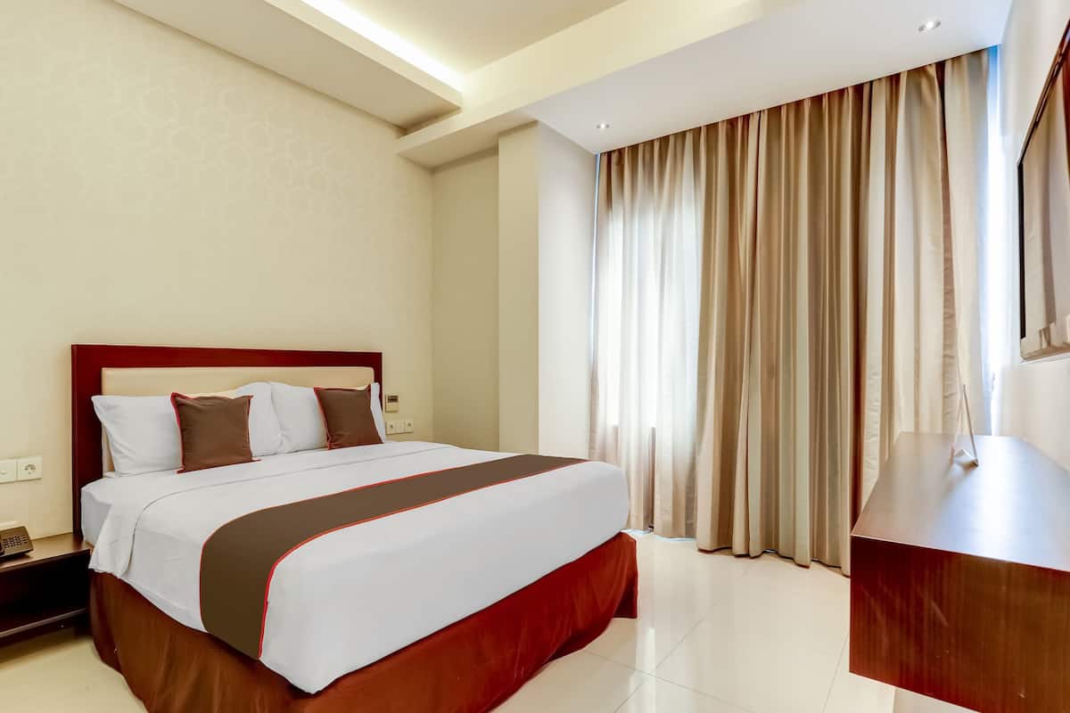 Habitació d'hotel amb un llit gran, una petita taula de nit de fusta, un escriptori i una finestra amb cortines.