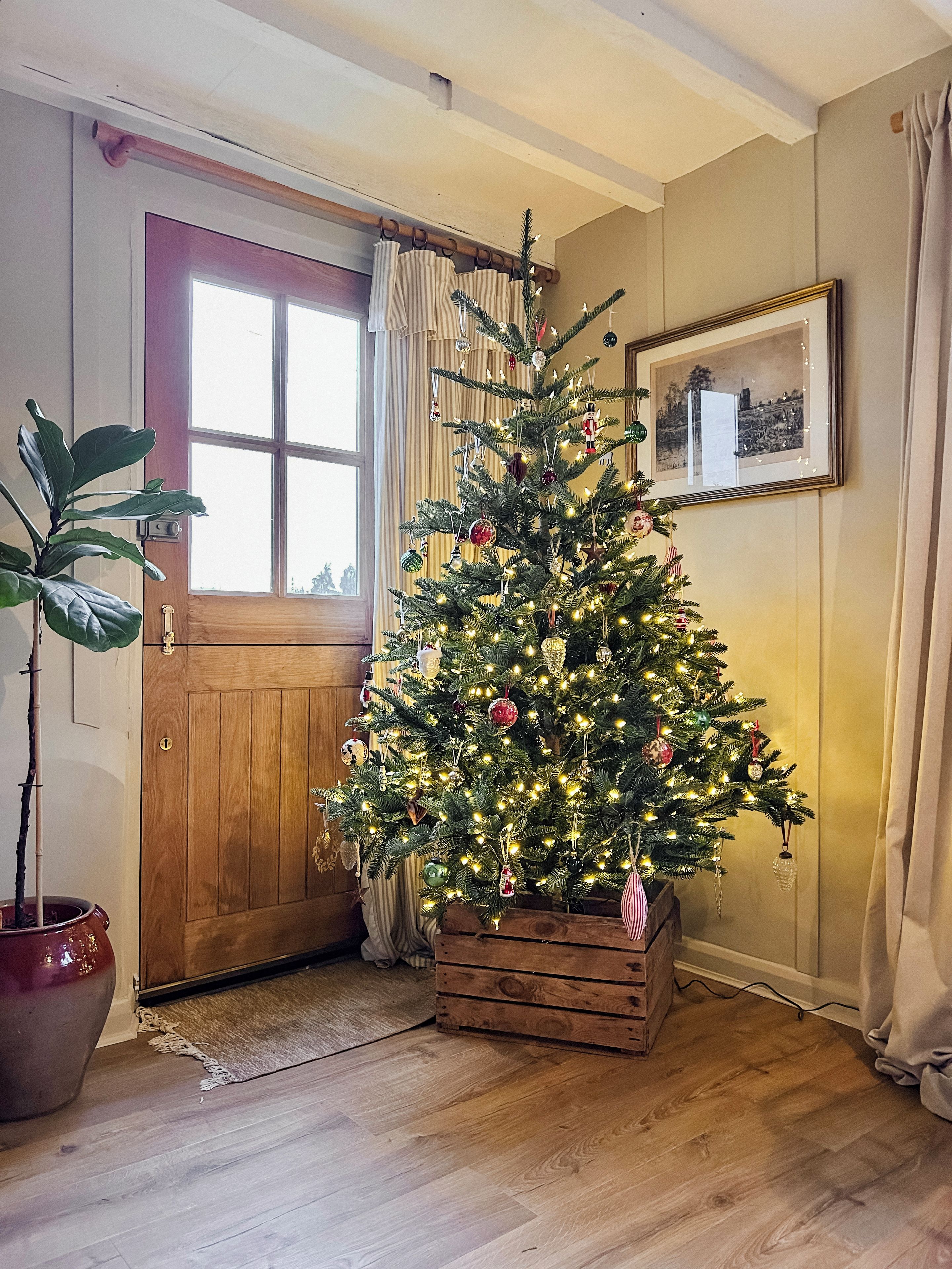 Un sapin de Noël placé dans un coin, avec une porte en bois et un tableau encadré accroché au mur.