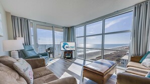 Amplia sala de estar con ventanas grandes que ofrecen vistas a la playa y al océano. Hay un letrero de Big Fish Rentals, un sofá y una mesita de centro.