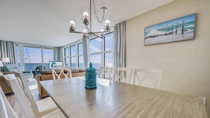 Amplia habitación con un gran comedor, sillas blancas, una araña y un cuadro de una playa.