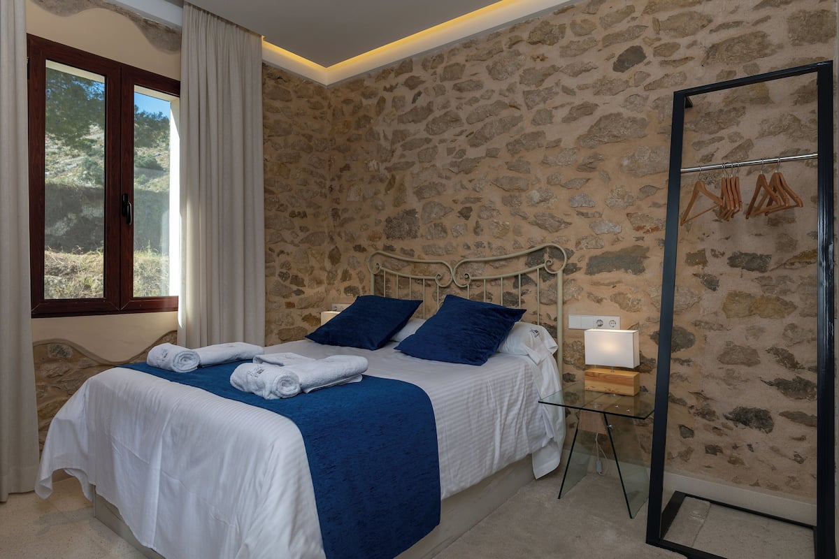 A bedroom with a stone wall, a bed with blue bedding, a bedside table with a lamp, and a window with curtains.