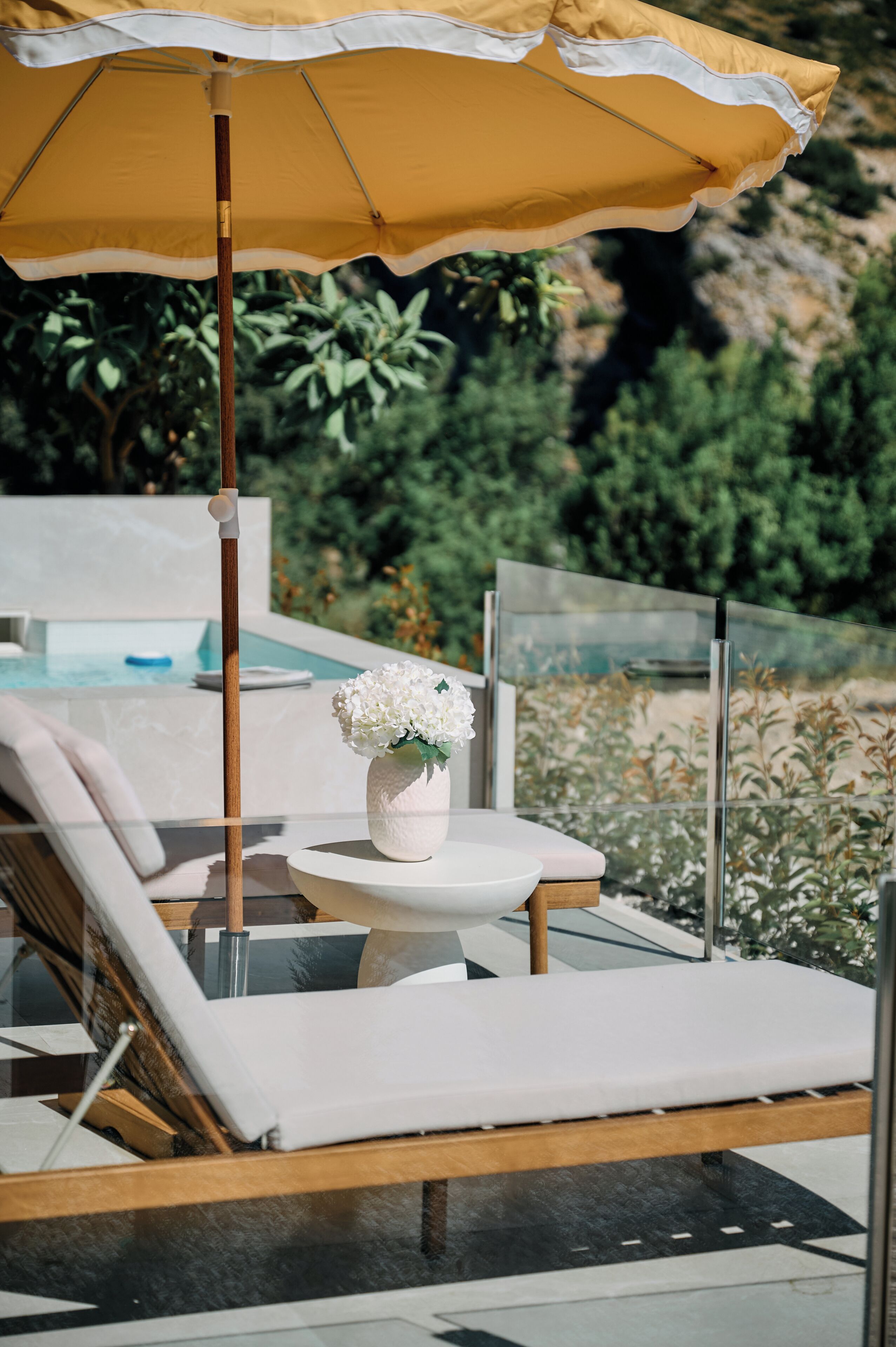 Ein Liegebereich am Pool mit einem gelben Sonnenschirm, einer weißen Vase mit Blumen und einer Glasreling.